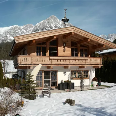 Ein charmantes Holzhaus mit Balkon, umgeben von verschneiten Wiesen. Im Hintergrund sind beeindruckende Berge zu sehen.