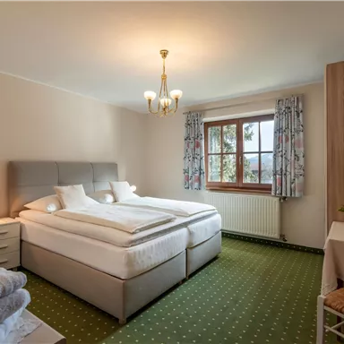 A cozy bedroom with two single beds and a large window. The room is bright and well-furnished, with green carpet flooring.