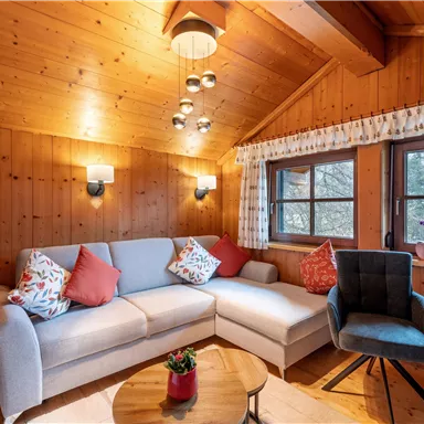A cozy living room with wood paneling and large windows. The furnishings consist of a comfortable sofa and a round table.