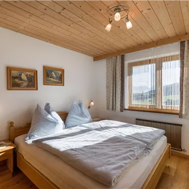 A cozy bedroom with a wooden ceiling and large windows. There is a double bed, two pillows, and decorative pictures on the wall.