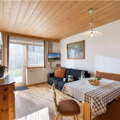 A cozy living space with wooden floors and a wooden ceiling. There is a dining area with a table and a couch, as well as large windows that let in a lot of light.