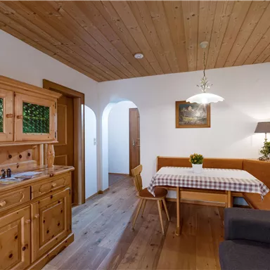 A cozy dining area with wooden furniture and a table covered with a checked tablecloth. The walls are bright, and the wooden ceiling gives the room a warm atmosphere.
