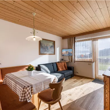 A cozy living room with a dining area, a dark blue couch, and large windows. The wooden floor and wooden ceiling give the room a warm atmosphere.