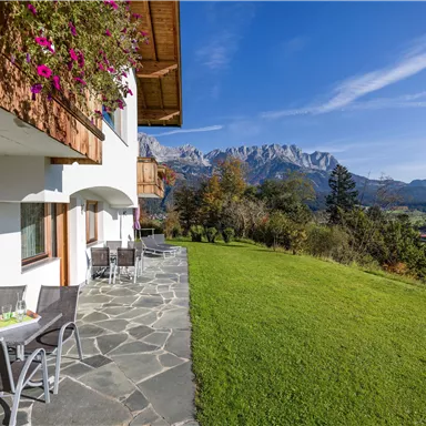 A beautiful view of the mountains from a garden with a lawn. On the terrace, there are tables and chairs to enjoy nature.
