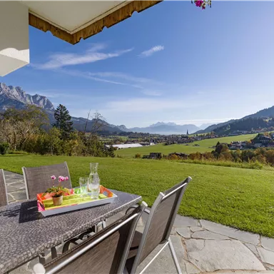 A terrace with modern furniture offers a view of the hills and mountains. The sky is clear and the surroundings are green and inviting.