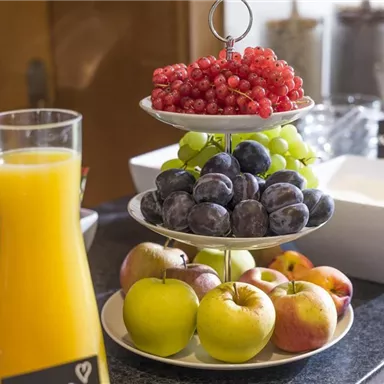 Ein dekorativer Obstteller mit roten Johannisbeeren, Trauben, Pflaumen und Äpfeln. Dazu stehen Gläser mit frisch gepresstem Orangensaft.
