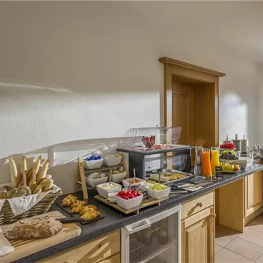 Eine einladende Frühstücksbar mit frischen Brötchen, Aufschnitt und Obst. Der Raum hat große Fenster, die einen Blick auf die umliegende Natur bieten.