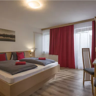 Ein gemütliches Hotelzimmer mit zwei Betten, modernen Möbeln und einem Schreibtisch. Große Fenster und rote Vorhänge sorgen für ein einladendes Ambiente.