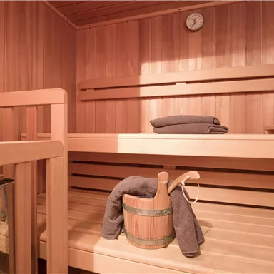 Ein gemütlicher Saunaraum mit Holzverkleidung. Im Inneren befinden sich Holzbänke, ein Handtuch und ein Eimer.