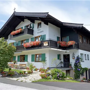 Ein charmantes Gebäude im alpinen Stil mit Blumenkästen und grünen Fensterläden. Der Eingang ist von gepflegten Pflanzen umgeben.