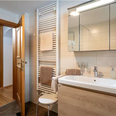 Ein modernes Badezimmer mit einer weißen Waschtischkombination und einem großen Spiegel. Im Hintergrund sind eine Holztür und ein Handtuchhalter zu sehen.