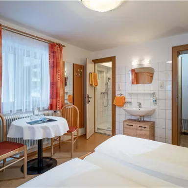 Ein gemütliches Gästezimmer mit zwei Einzelbetten, einem Tisch und Stühlen. Das Zimmer hat große Fenster mit Vorhängen und ein eigenes Bad.