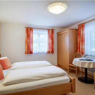 Ein gemütliches Zimmer mit zwei Einzelbetten und einem kleinen Tisch. Die Fenster sind hell und mit Vorhängen dekoriert.