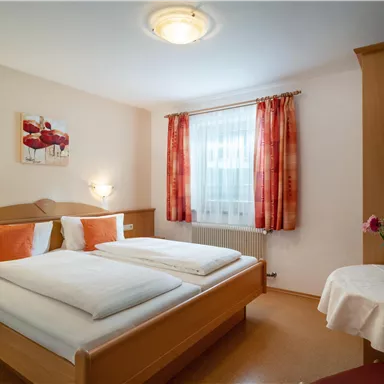 Ein gemütliches Schlafzimmer mit zwei Betten, hellen Wänden und warmen Farben. Neben einem Tisch mit einer Blumenvase gibt es große Fenster mit Vorhängen.