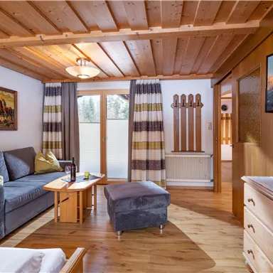 Ein gemütlicher Wohnraum mit rustikalem Holzdekor, einem Sofa und einem TV. Große Fenster sorgen für viel Tageslicht und bieten einen Blick nach draußen.