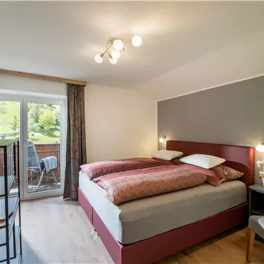 A cozy bedroom with a large bed and modern decor. The windows offer a view of greenery and let in plenty of light.