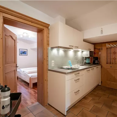 A modern kitchen with light cabinets and a countertop. In the background, a bedroom with a double bed can be seen.