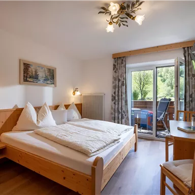 A cozy room with a double bed, bright walls, and wood paneling. There is a small table and chairs as well as a balcony with a view of the greenery.
