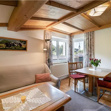 A cozy living room with a gray sofa and a wooden table. The windows offer a view of nature, and fresh flowers are on the table.