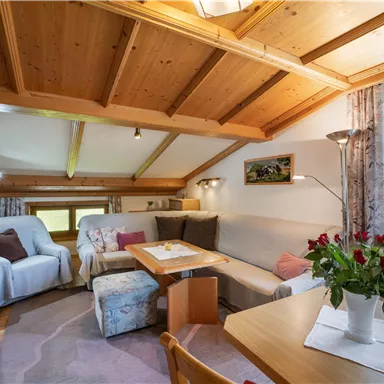 A cozy living room with wooden ceilings and a large window. There is a seating area, a table, and a television.