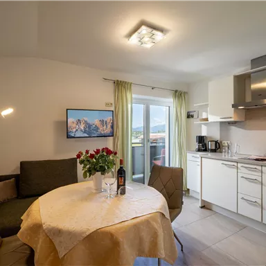 A modern living room with a kitchen and a dining table. On the table, there are roses and a bottle of wine, while light shines from a lamp in the room.