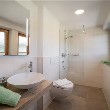 A modern bathroom with a shower and a large mirror. The walls are bright, and there are green towels on a wooden shelf.