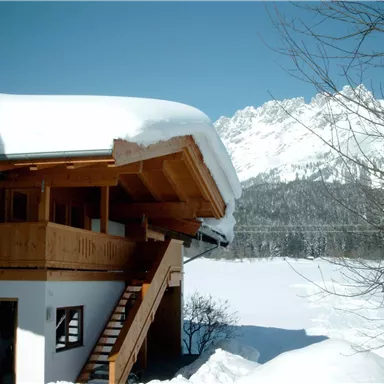 A beautiful wooden house in the snow with a sloping roof. In the background, impressive mountains and a blue sky can be seen.