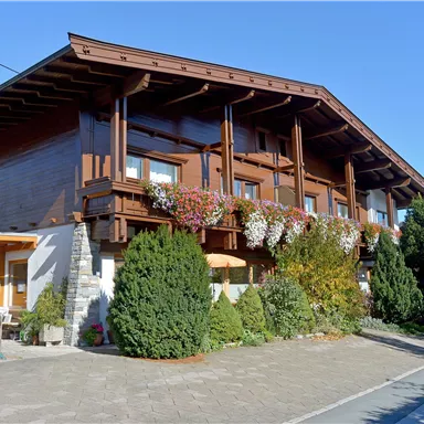 Ein schönes Holzhaus mit blühenden Balkonpflanzen. Umgeben von Bäumen und einer ruhigen Straße.