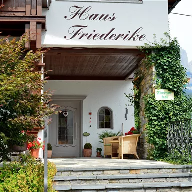 Ein einladendes Haus mit dem Namen "Haus Friedrike", umgeben von Pflanzen und Bäumen. Im Hintergrund sind Berge zu sehen.