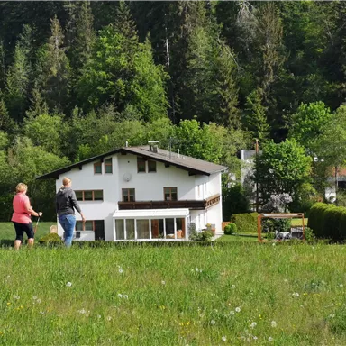 Ein gemütliches Haus umgeben von einer grünen Wiese und Bäumen. Zwei Personen spazieren entspannt am Haus vorbei.