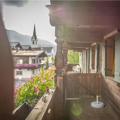 Ein Balkon mit Holzelementen und blühenden Pflanzen. Im Hintergrund sieht man eine kleine Kirche und die Berge.
