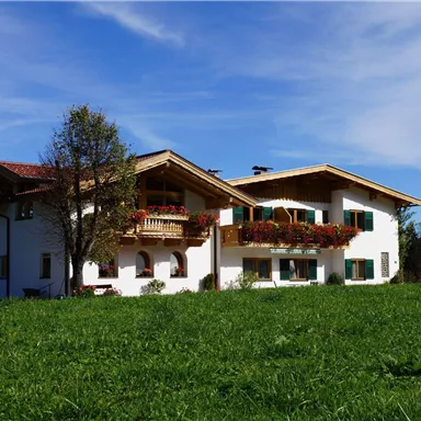 Ein schönes Landhaus mit einem großen Garten und bunten Blumenbalkonen. Im Hintergrund sind sanfte Hügel und ein klarer blauer Himmel sichtbar.