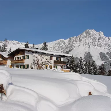 Ein schönes Haus in einer schneebedeckten Landschaft mit Bergen im Hintergrund. Der Himmel ist klar und blau.