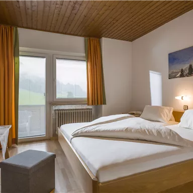 Ein modernes Schlafzimmer mit einem Doppelbett und einer gemütlichen Holzdecke. Große Fenster lassen viel Licht herein und bieten einen Blick ins Grüne.