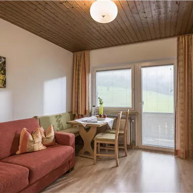 Ein gemütlicher Wohnraum mit einem roten Sofa und einem Esstisch. Die großen Fenster bieten eine Aussicht auf die grüne Landschaft.