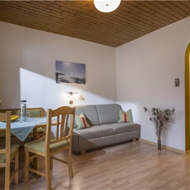 Ein gemütlicher Wohnraum mit einem Tisch und Stühlen sowie einem Sofa. Eine warme Atmosphäre mit Holzdecken und einem kleinen Fenster, das Licht hereinlässt.