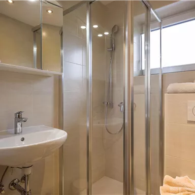 Ein modernes Badezimmer mit einer Duschkabine und einem Waschbecken. Helle Fliesen und ein Fenster sorgen für eine freundliche Atmosphäre.