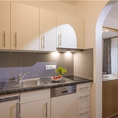 Eine moderne Küche mit hellen Schränken und einer Arbeitsfläche. Im Hintergrund ist ein gemütliches Schlafzimmer sichtbar.