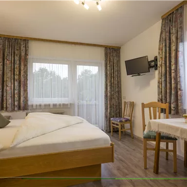 Ein gemütliches Schlafzimmer mit einem Doppelbett und einem Tisch. Helle Vorhänge und eine angenehme Atmosphäre machen den Raum einladend.