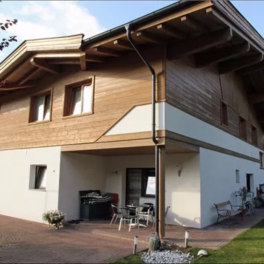 Ein modernes Haus mit Holzverkleidung und einer breiten Terrasse. Der Garten ist mit Sitzgelegenheiten und Pflanzen gestaltet.