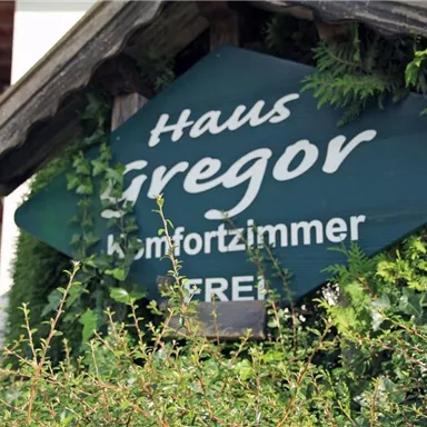 Ein Schild mit der Aufschrift „Haus Gregor“ und „Komfortzimmer frei“ ist von Pflanzen umgeben. Im Hintergrund sind Elemente eines traditionellen Gebäudes sichtbar.