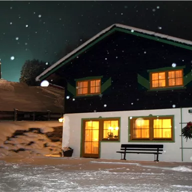 Ein hübsches, dunkelblaues Haus im Schnee mit beleuchteten Fenstern. Im Hintergrund sieht man sanfte, verschneite Hügel und einen beleuchteten Turm.