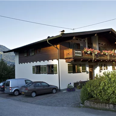Ein traditionelles Haus in den Bergen mit einem Balkon voller Blumen. Parkplätze sind vor dem Gebäude vorhanden und die Umgebung ist grün und einladend.