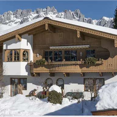 A beautiful wooden house with balconies, surrounded by snow. In the background, majestic mountains rise under a clear blue sky.
