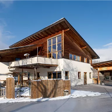 Ein modernes Haus mit Holzverkleidung und großen Fenstern. Draußen liegt Schnee und der Himmel ist blau.