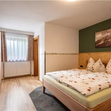 Ein gemütliches Schlafzimmer mit einem großen Bett und kühlem Holzmobiliar. Der Raum hat große Fenster und ein dekoratives Bild an der Wand.