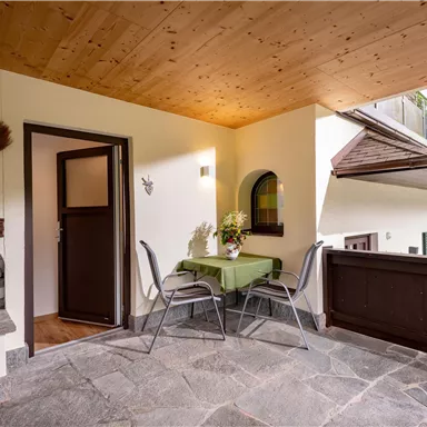 Ein gemütlicher Balkon mit einem Tisch und zwei Stühlen. Im Hintergrund sind ein Kamin und die Natur zu sehen.