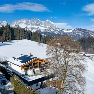 A picturesque winter landscape with snow-covered mountains and a wooden house. In the foreground are green trees and gentle hills.