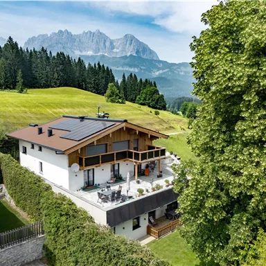 Ein modernes Haus umgeben von üppigem Grün und Bäumen. Im Hintergrund erheben sich majestätische Berge unter einem klaren Himmel.