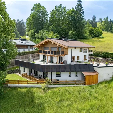 Ein modernes Haus in einer grünen Landschaft mit Bäumen und Wiesen. Die Architektur zeigt Balkone und eine gemütliche Terrasse.